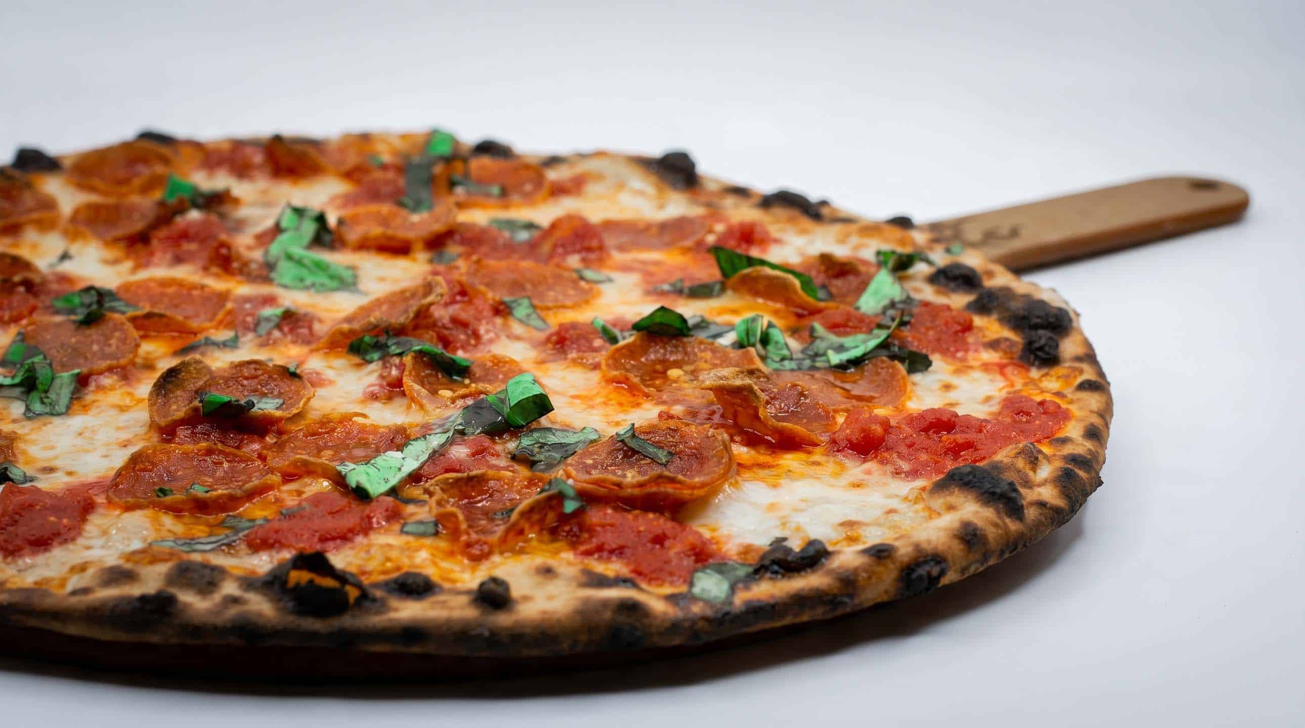 Wood-fired pepperoni pizza topped with fresh basil on a wooden peel